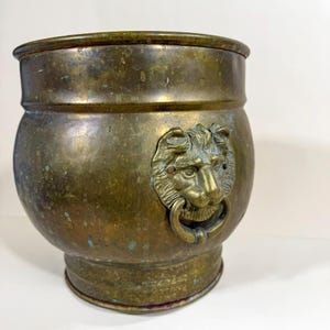May include: A weathered brass planter with a lion's head detail. The planter has a rounded body and a flared base. The lion's head features a ring for a handle. The brass has a patina with green and blue spots.