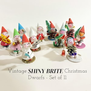 May include: A collection of eleven vintage Shiny Brite Christmas dwarf figurines. Each dwarf has a painted face, a pointed hat, and a detailed outfit. Some are holding musical instruments or small Christmas trees. The figurines are on small, round bases.