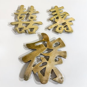May include: Three brass decorative wall plaques featuring cut-out Chinese characters. The top left plaque displays the double happiness symbol, the top right shows the character for longevity, and the larger bottom plaque features the character for good fortune.
