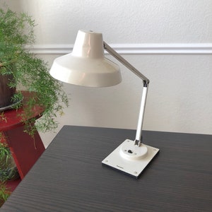May include: A white desk lamp with a chrome arm and a white base. The lamp has a white shade and a black switch on the base. The lamp is on a dark wood surface.