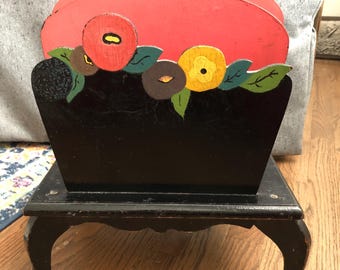 Puede incluir: Un revistero de madera negro con la parte superior roja y un diseño floral pintado. El revistero tiene cuatro patas y una base decorativa.