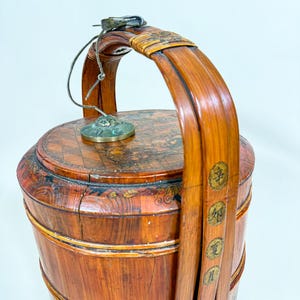 May include: A vintage wooden bucket with a curved handle and a lid. The bucket is made of reddish-brown wood with decorative elements and Chinese characters. A metal clasp and a small brass disc are attached to the lid.