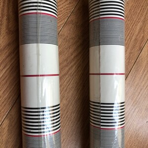 May include: Two rolls of black and white striped wallpaper with a thin red stripe. The wallpaper is packaged in clear plastic.