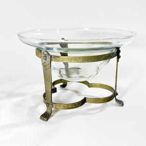 May include: A clear glass bowl with a wide rim, supported by an ornate brass stand. The stand features decorative floral patterns and a three-legged base, creating an elegant, vintage aesthetic.