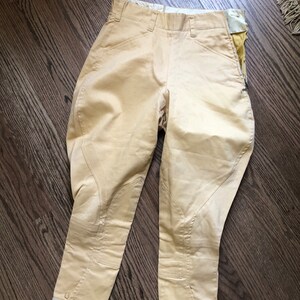 Op de afbeelding: Een paar beige rijbroeken met witte bies aan de onderkant. De broek heeft een ritszak aan de rechterkant.