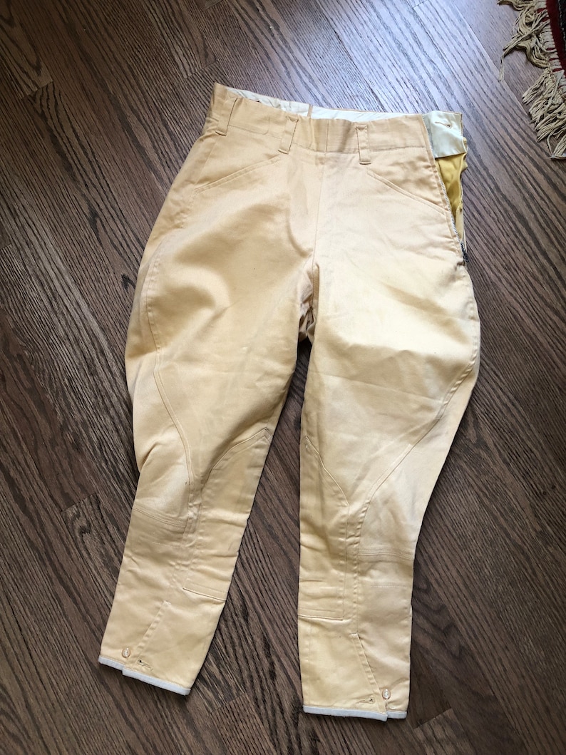 Op de afbeelding: Een paar beige rijbroeken met witte bies aan de onderkant. De broek heeft een ritszak aan de rechterkant.
