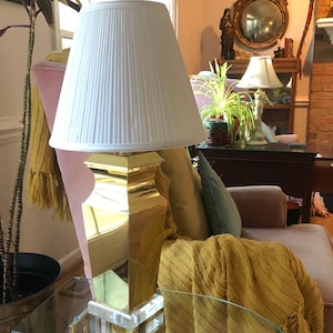 May include: A gold table lamp with a white pleated lampshade sits on a glass table. The lamp is in a living room setting with a pink armchair and a yellow throw blanket.