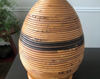 May include: A woven rattan lamp base with a natural brown finish and a dark brown band around the middle. The base is shaped like an egg and sits on a wooden pedestal.