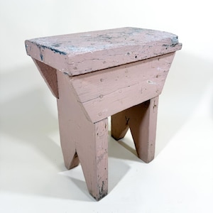 May include: A small, weathered, pink wooden stool with a rectangular top and angled legs. The stool has visible nail heads and a distressed finish, suggesting age and use. The overall design is simple and rustic.