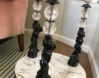 May include: A round side table with a clear glass top and a white faux marble lower shelf. The table features three legs, each designed as a black cherub figure supporting two stacked clear faceted crystal spheres beneath the glass top.
