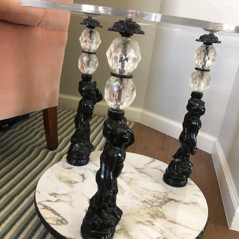 May include: A round glass table with a white marble top and three black cherub-shaped legs. Each leg is topped with a crystal ball and a smaller crystal orb.