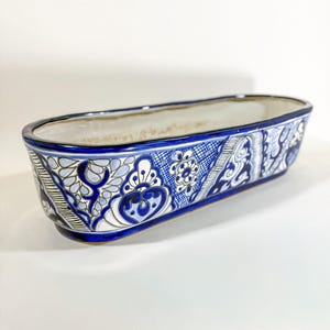 May include: An elongated oval ceramic planter featuring a hand-painted blue and white floral pattern. The exterior displays intricate scrollwork, geometric lines, and botanical motifs in varying shades of blue, while the interior is a solid off-white.