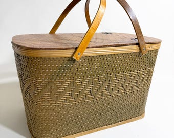 May include: A vintage picnic basket with a woven olive green and tan design. The basket has a wooden lid and two arched wooden handles. The basket is rectangular with rounded edges and a decorative woven pattern.