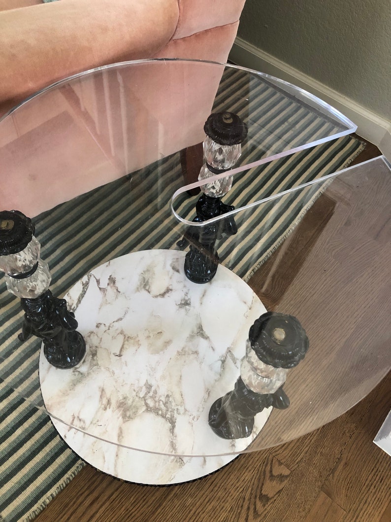 May include: A round, clear glass table with a white marble top. The table has three black and gold legs with decorative finials.