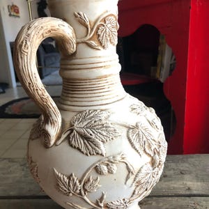 May include: A white ceramic pitcher with a detailed design of leaves and vines. The pitcher has a wide base and a rounded body. The handle is shaped like a tree branch.