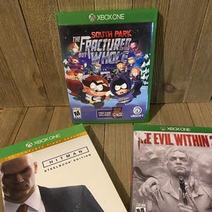 May include: Three Xbox One video game cases. The first case is for South Park: The Fractured But Whole, the second case is for Hitman: The Complete First Season Steelbook Edition, and the third case is for The Evil Within.