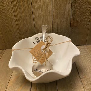 May include: A white ceramic bowl with a wavy rim and a silver-toned metal spoon resting inside. The bowl features the text "Nachos ordinarios" and the spoon is engraved with "MEX-ELLENT". A brown paper tag is tied to the spoon with twine.