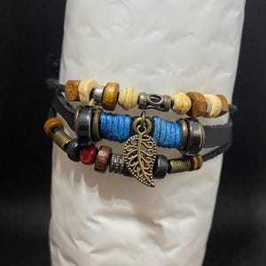 May include: A multi-strand bracelet featuring a combination of wooden, metallic, and blue cord beads. The design includes a central bronze-toned metal leaf charm, dark leather-like straps, and various cylindrical spacers in silver and brass tones.