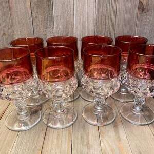 May include: A set of eight drinking glasses featuring a deep red upper bowl and a clear, textured glass base with a repeating circular pattern. Each glass has a short, sturdy stem and a wide, flat circular foot, designed for serving beverages.