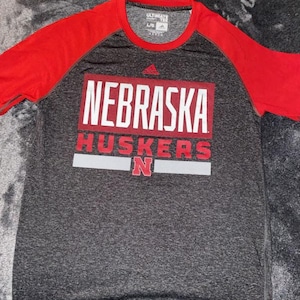 May include: A gray and red Adidas t-shirt featuring the text "NEBRASKA HUSKERS" in white and red block letters. The shirt has red sleeves and a crew neck. The Adidas logo is visible.