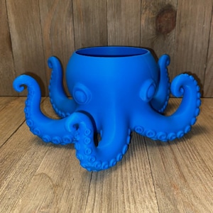 May include: A bright blue octopus-shaped planter featuring a smooth, matte finish. The vessel has a wide, circular opening at the top and eight curved tentacles extending outward from the base, each detailed with small, raised circular suction cups.