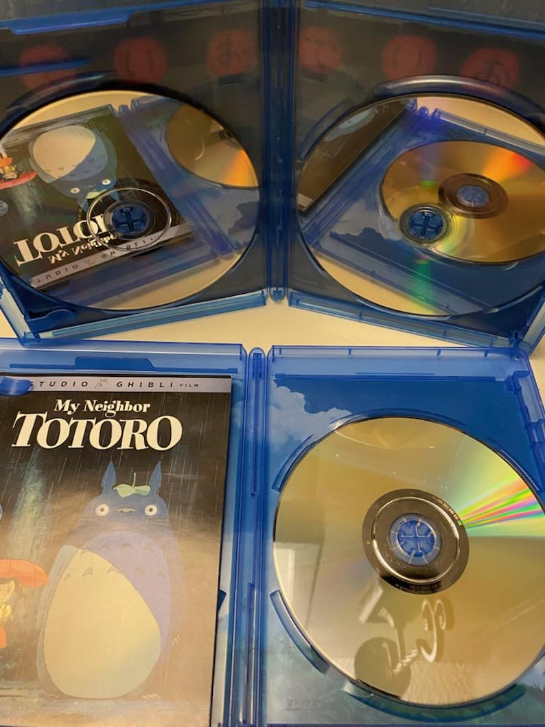 May include: Blu-ray disc set of the animated film "My Neighbor Totoro" in a blue case. The cover art features the film's title and a depiction of Totoro. The discs are gold and reflect light.