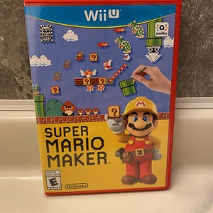 May include: A red and blue Wii U game case for Super Mario Maker. The cover features Mario in a construction hat, pixelated graphics, and the Wii U logo. The game is rated E for Everyone.