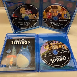 May include: Four Blu-ray cases are open, displaying the discs for "Spirited Away" and "My Neighbor Totoro." The cases are blue, and the discs feature colorful illustrations from the animated films. The "My Neighbor Totoro" case shows the film's title.