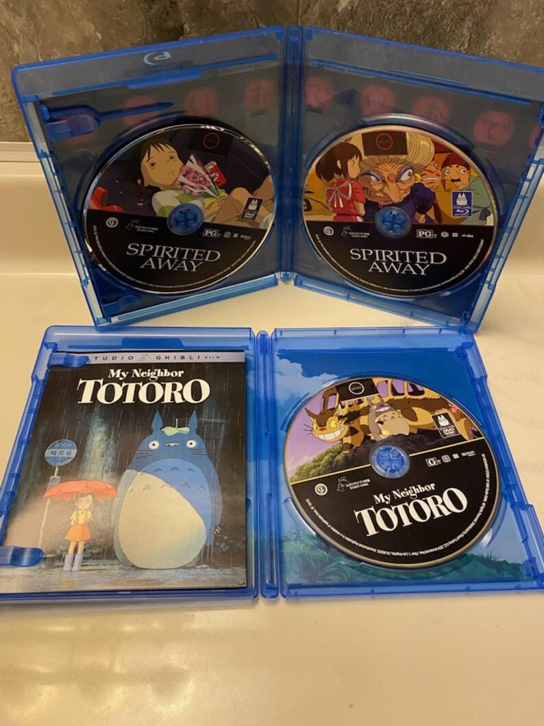 May include: Four Blu-ray cases are open, displaying the discs for "Spirited Away" and "My Neighbor Totoro." The cases are blue, and the discs feature colorful illustrations from the animated films. The "My Neighbor Totoro" case shows the film's title.