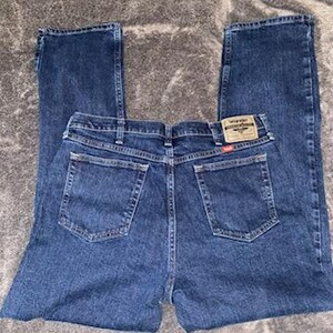 May include: Dark blue denim jeans with a classic five-pocket design. The jeans feature a straight leg cut and a brown and red Wrangler brand patch on the back pocket. The jeans are laid flat on a gray surface.