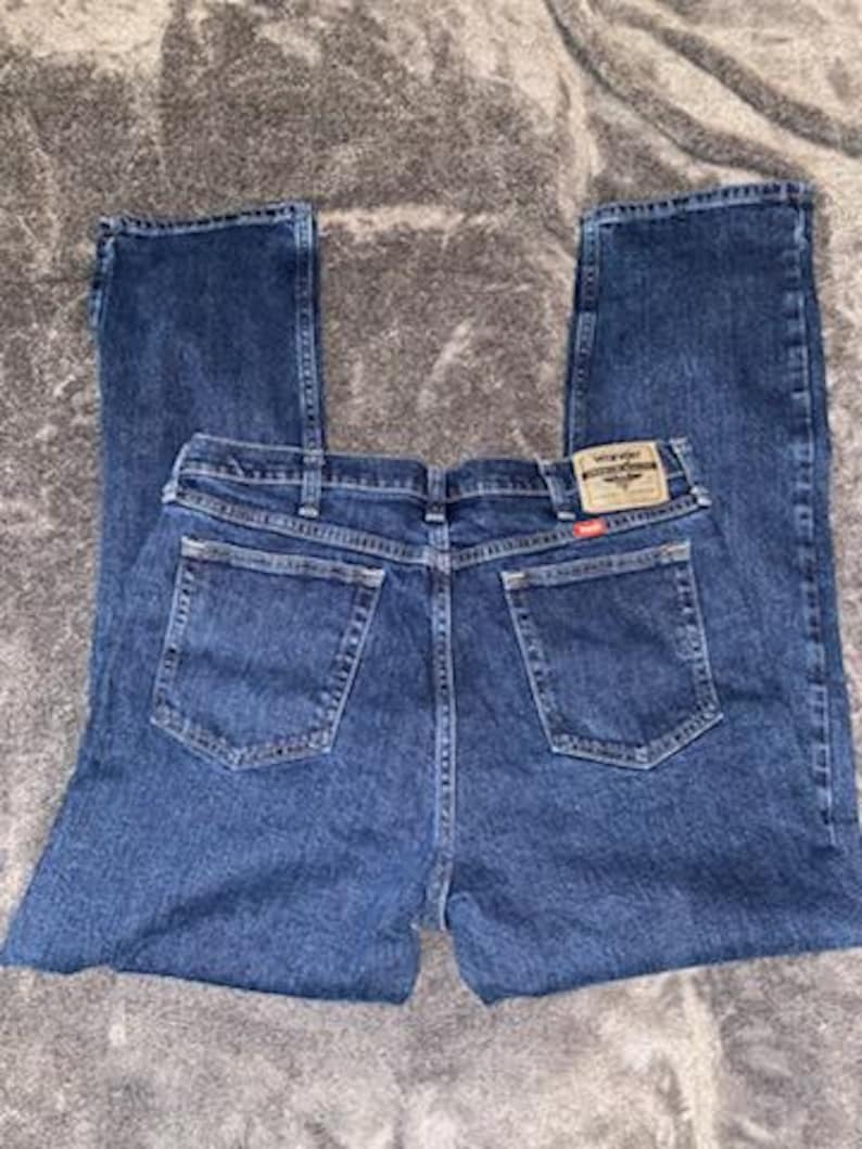 May include: Dark blue denim jeans with a classic five-pocket design. The jeans feature a straight leg cut and a brown and red Wrangler brand patch on the back pocket. The jeans are laid flat on a gray surface.