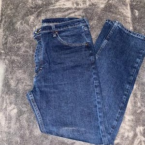 May include: Dark blue denim jeans. The jeans have a classic five-pocket design, a button and zipper closure, and belt loops. The denim has a slightly faded appearance, with subtle wear marks.