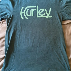 May include: A teal-colored short-sleeved t-shirt with the word "Hurley" printed across the chest in a lighter green. The shirt has a crew neck and appears to be made of a soft, comfortable material. The Hurley logo is stylized.