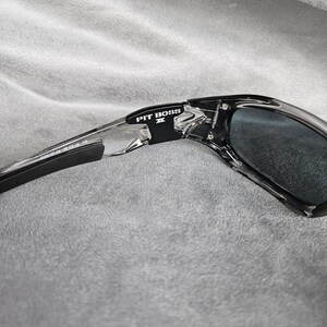 May include: Black and clear sunglasses with the text "PIT BOSS II" on the frame. The sunglasses have dark lenses and a sleek, modern design. The frame is a combination of black and clear plastic.