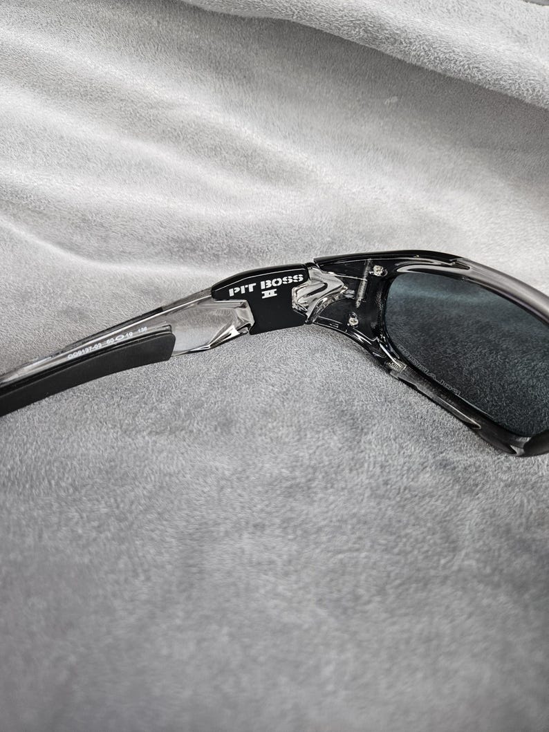 May include: Black and clear sunglasses with the text "PIT BOSS II" on the frame. The sunglasses have dark lenses and a sleek, modern design. The frame is a combination of black and clear plastic.
