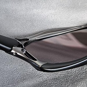 Puede incluir: Primer plano de gafas de sol negras con un dise&ntilde;o elegante y moderno. Las monturas son negras con detalles plateados y las lentes son de color ahumado oscuro. Las gafas de sol tienen una forma curvada.