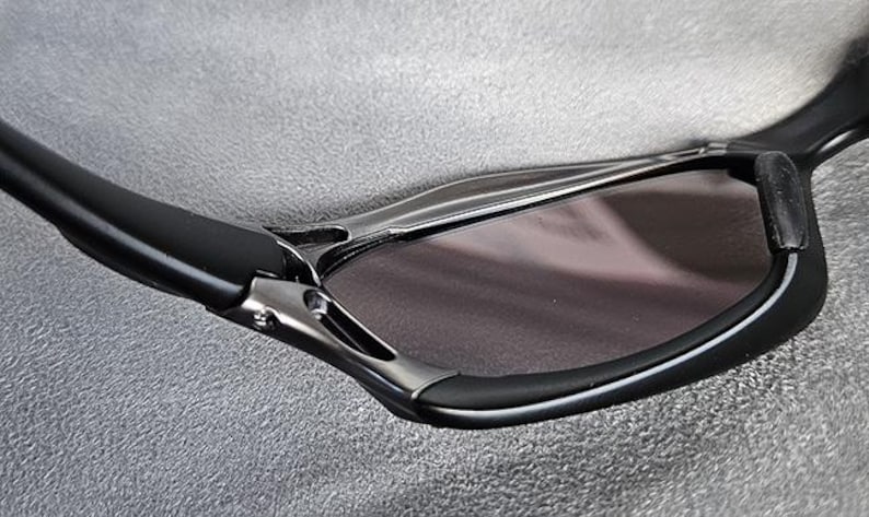 Puede incluir: Primer plano de gafas de sol negras con un dise&ntilde;o elegante y moderno. Las monturas son negras con detalles plateados y las lentes son de color ahumado oscuro. Las gafas de sol tienen una forma curvada.