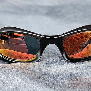 Puede incluir: Gafas de sol negras con lentes espejadas naranjas y amarillas. Las gafas tienen un dise&ntilde;o curvo y envolvente. Las lentes reflejan el entorno. Sobre un fondo gris.
