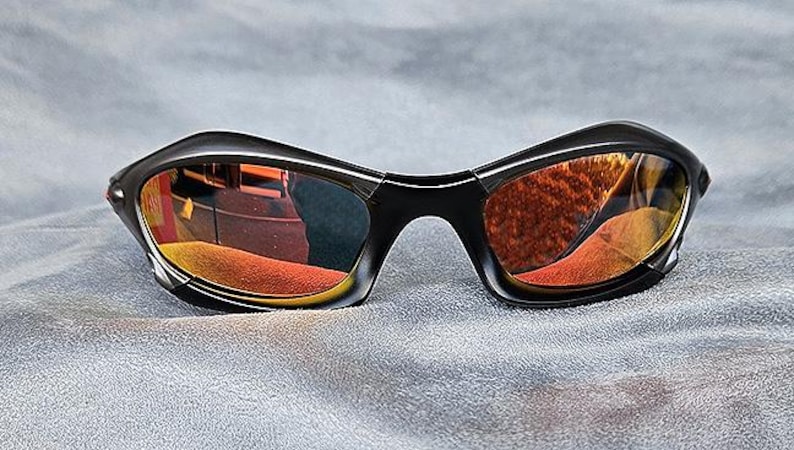 Puede incluir: Gafas de sol negras con lentes espejadas naranjas y amarillas. Las gafas tienen un dise&ntilde;o curvo y envolvente. Las lentes reflejan el entorno. Sobre un fondo gris.