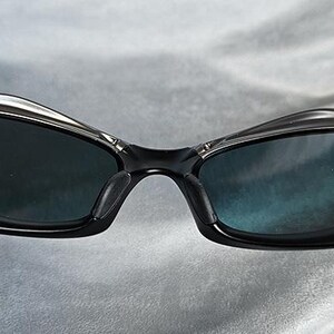 Puede incluir: Primer plano de gafas de sol negras con un dise&ntilde;o elegante y moderno. Las gafas de sol tienen lentes oscuras y una montura negra con detalles plateados. La imagen est&aacute; bien iluminada, destacando los detalles de las gafas.