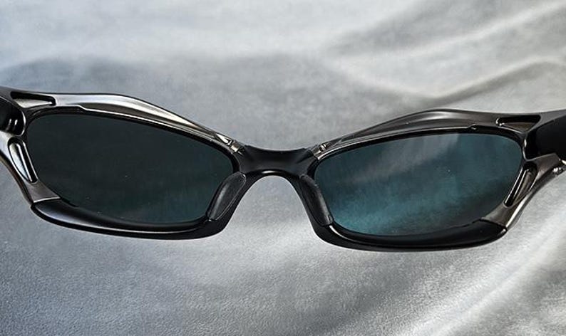 Puede incluir: Primer plano de gafas de sol negras con un dise&ntilde;o elegante y moderno. Las gafas de sol tienen lentes oscuras y una montura negra con detalles plateados. La imagen est&aacute; bien iluminada, destacando los detalles de las gafas.