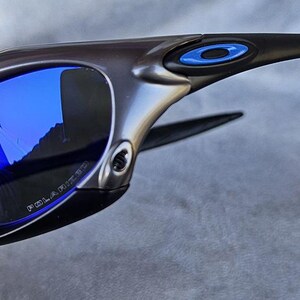 Puede incluir: Gafas de sol gris oscuro con lentes azules espejadas. Las gafas tienen un dise&ntilde;o elegante y moderno con montura negra y detalles azules. Las lentes est&aacute;n polarizadas. La palabra "POLARIZED" est&aacute; impresa en la lente.