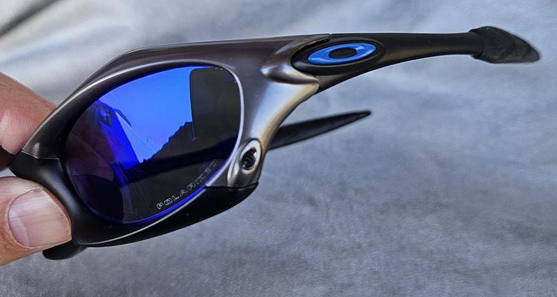 Puede incluir: Gafas de sol gris oscuro con lentes azules espejadas. Las gafas tienen un dise&ntilde;o elegante y moderno con montura negra y detalles azules. Las lentes est&aacute;n polarizadas. La palabra "POLARIZED" est&aacute; impresa en la lente.
