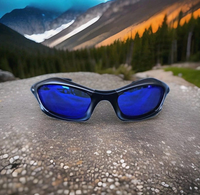 Puede incluir: Gafas de sol negras con lentes azules reflectantes sobre una roca texturizada. Las gafas de sol tienen un dise&ntilde;o deportivo, con una montura curvada y un aspecto elegante y moderno.