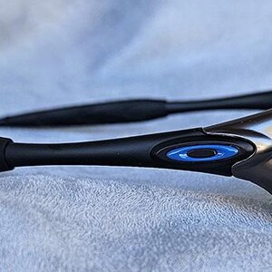 Puede incluir: Gafas de sol deportivas negras y plateadas con lentes azules. Las gafas de sol tienen un logotipo ovalado azul en la patilla y patillas negras texturizadas. La montura tiene un acento plateado.
