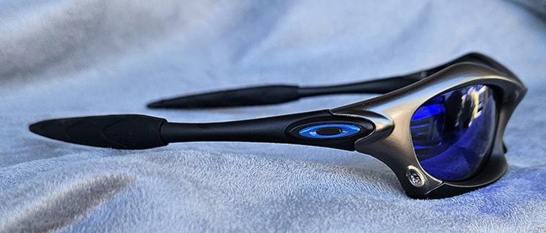 Puede incluir: Gafas de sol deportivas negras y plateadas con lentes azules. Las gafas de sol tienen un logotipo ovalado azul en la patilla y patillas negras texturizadas. La montura tiene un acento plateado.