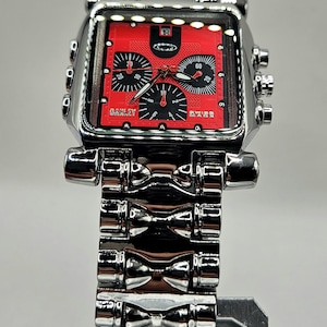 May include: A silver-toned Oakley watch with a red face and black accents. The watch features a square face with multiple dials and a stainless steel band. The watch is displayed on a silver stand.