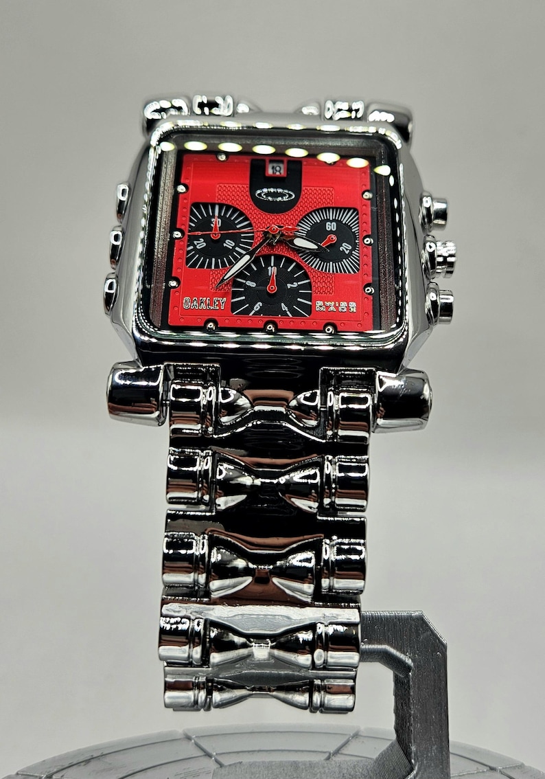 May include: A silver-toned Oakley watch with a red face and black accents. The watch features a square face with multiple dials and a stainless steel band. The watch is displayed on a silver stand.