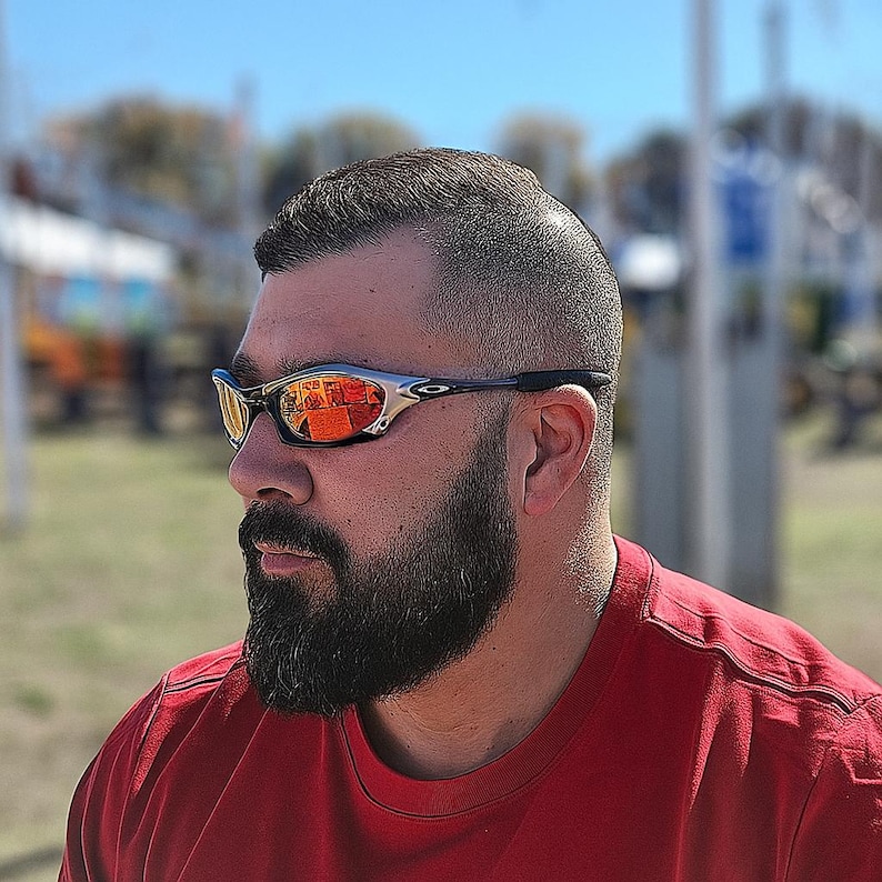 Może przedstawiać: Srebrne i czarne okulary przeciwsłoneczne z pomarańczowymi lustrzanymi soczewkami. Okulary mają sportowy design i są noszone przez osobę z brodą i czerwoną koszulką. Tło jest rozmyte.
