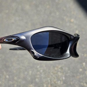 Peut inclure: Lunettes de soleil grises avec des verres fonc&eacute;s. Les lunettes ont une monture incurv&eacute;e et un petit logo argent&eacute; sur la branche. Les verres sont sombres et prot&egrave;gent du soleil.